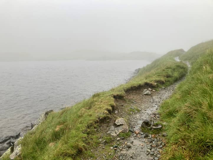 Weg führt an einem See entlang, es ist neblig und regnerisch, die Sicht ist schlecht