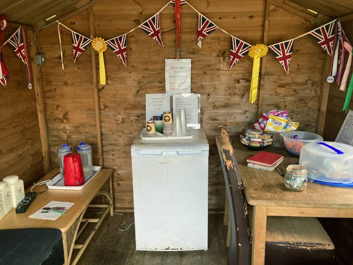 Blick in das Holzhäuschen. Ein Kühlschrank, ein Tisch mit Kuchen und Snacks. Kleine britische Flaggen schmücken den Raum