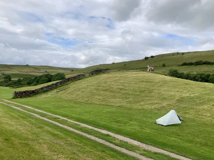 Aus erhöhter Position Blick auf grüne Hügel mit einem einzelnes weißes Zelt, davor führt ein Weg vorbei