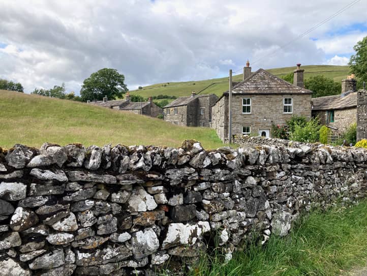 eine alte Steinmauer, dahinter zwischen den Hügeln ein kleines Dörfchen, die Häuser sind ebenfalls aus grauem Stein gebaut